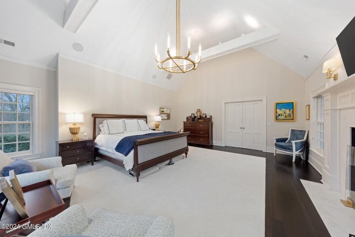 A bedroom with a bed, vaulted ceiling, window, chandelier, TV, chair, cabinet, and nightstand.