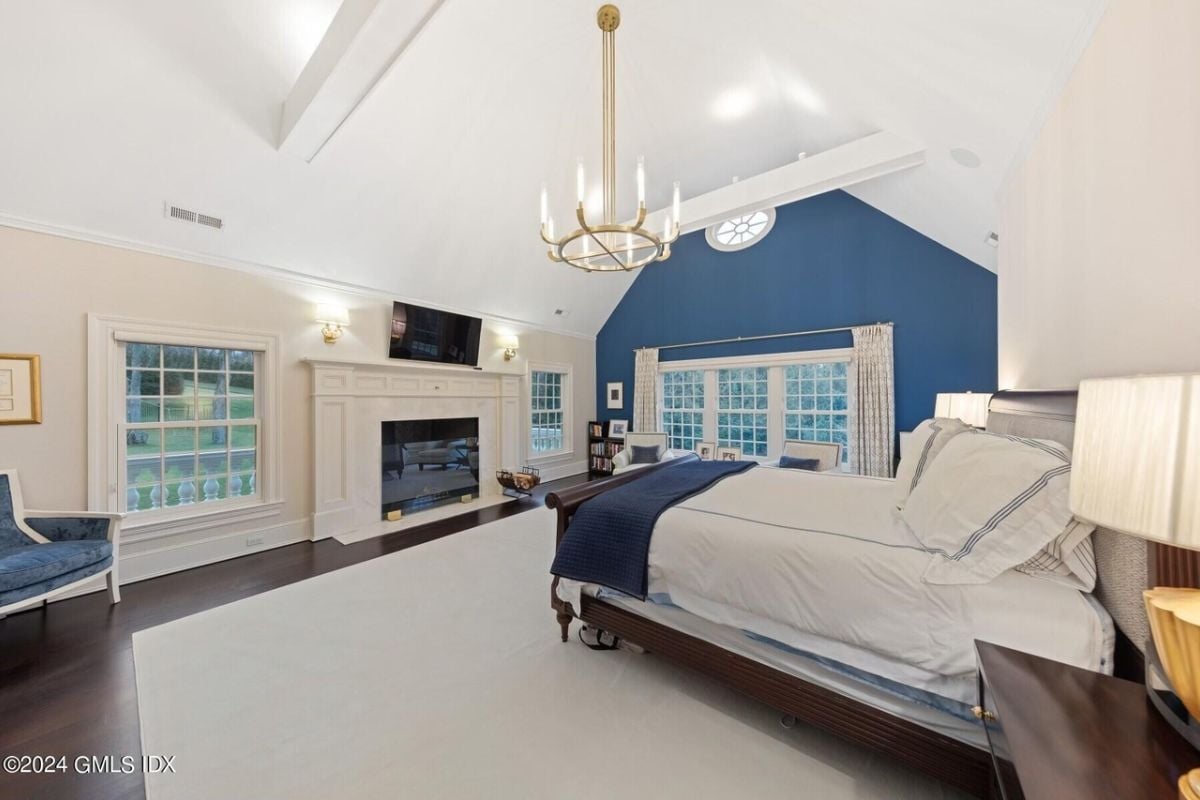A bedroom with a bed, vaulted ceiling, window, chandelier, TV, chair, firepllace, and nightstand.