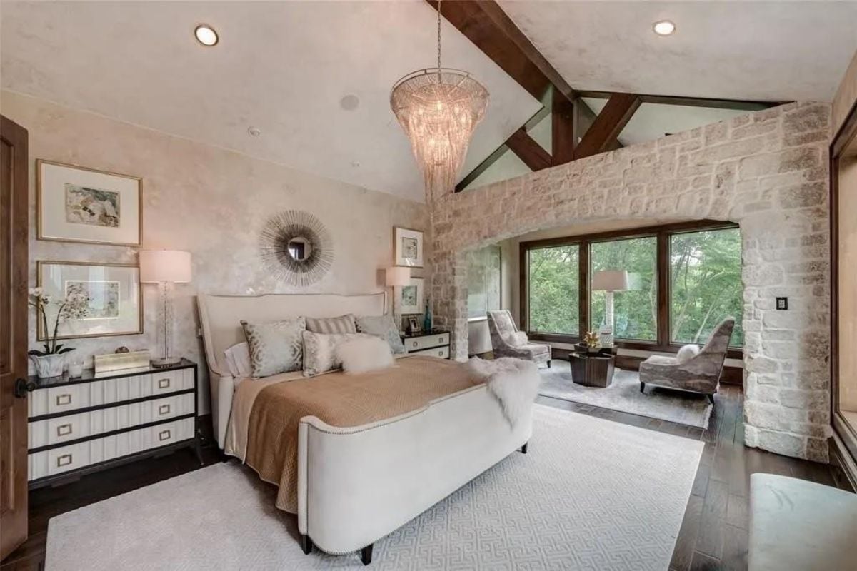 A bedroom with a vaulted ceiling, bed, cabinet, chandelier, windows, chairs, and coffee table.