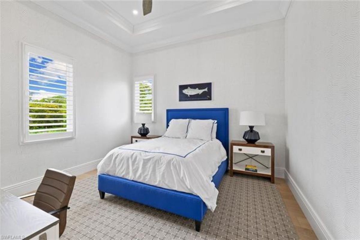 A bedroom with a bed, windows, a nightstand, a rug, a desk, and a chair.
