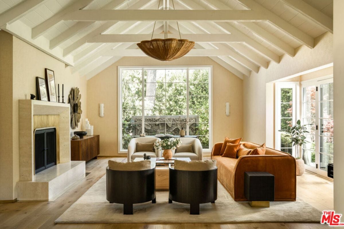 A living room with an exposed beam ceiling, fireplace, windows, couches, chairs, and a center table.