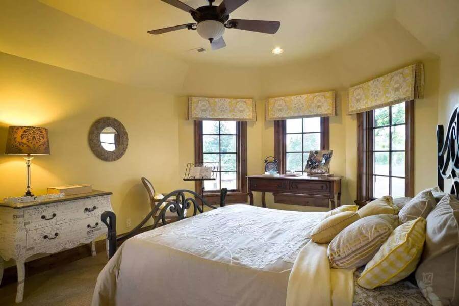 Bedroom with an ornate iron bed, a distressed dresser, and a dark wood desk against the bay window.