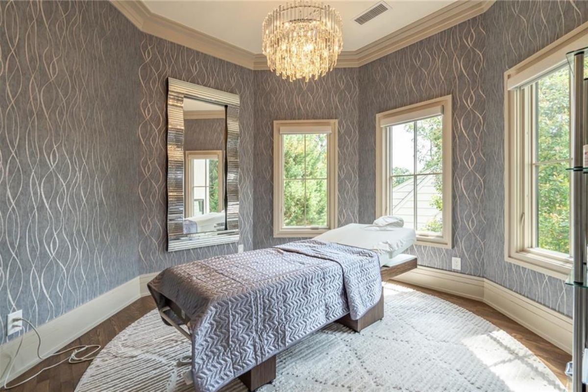 A massage room with windows, wallpaper, a chandelier, and a massage bed.