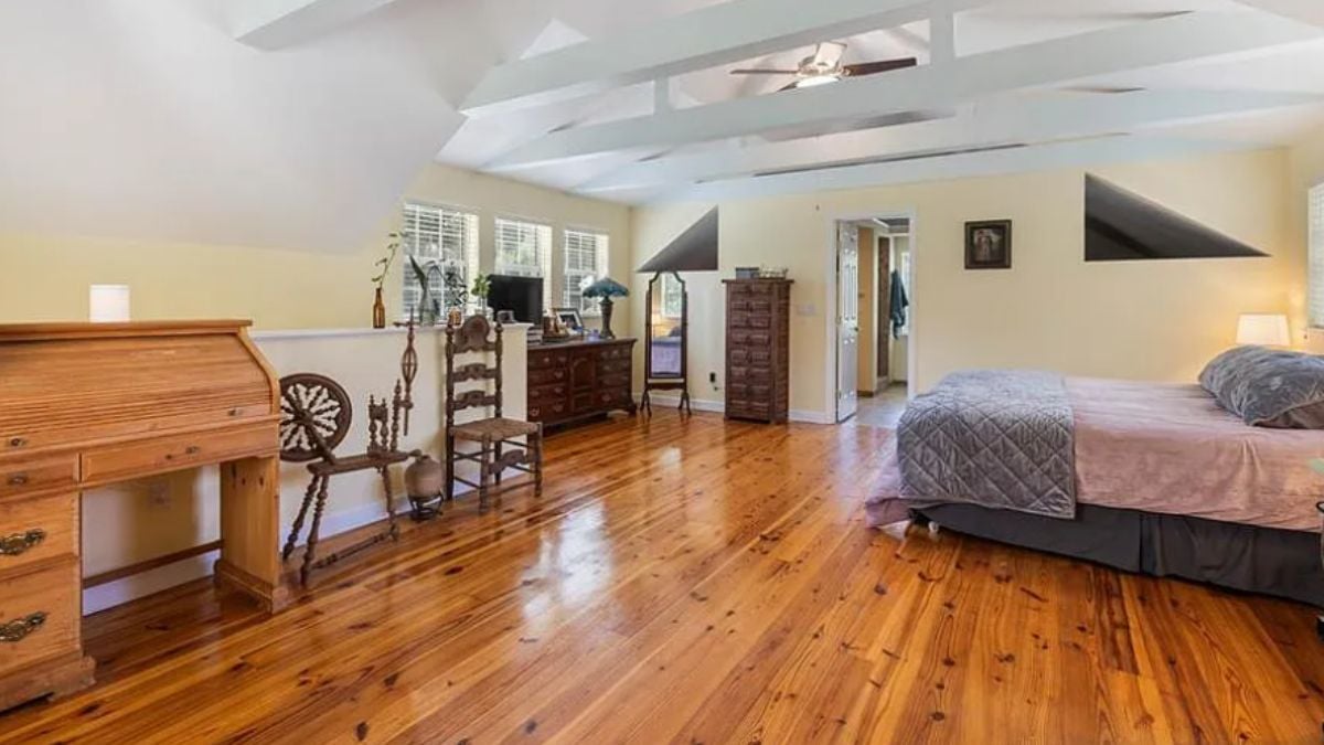 A bedroom with a bed, ceiling fan, exposed beam ceiling, windows, a cabinets, a nightstand, and a chair.