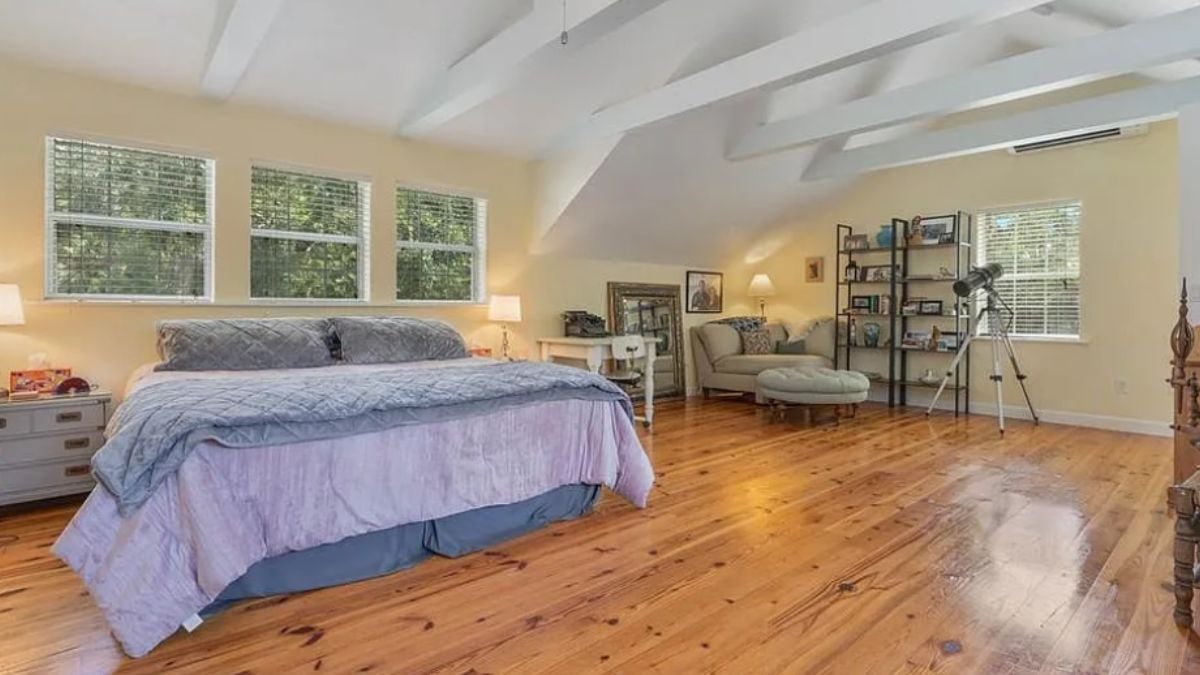 A bedroom with a bed, exposed beam ceiling, windows, a cabinet, a nightstand, and a desk with a chair.