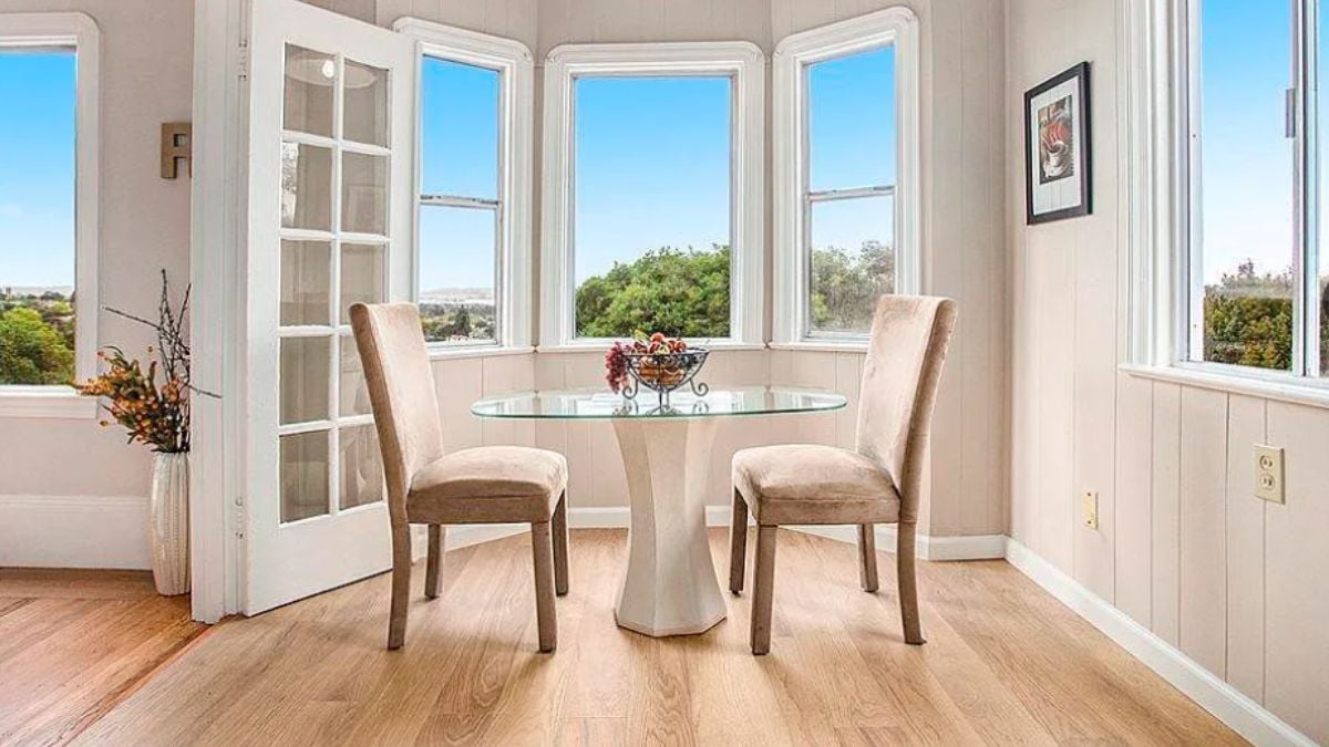 A breakfast nook with windows and a round glass table surrounded by chairs.