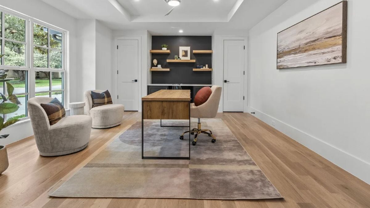 A home office with a painting, a shelf, windows, a rug, chairs, and a desk with an office chair.