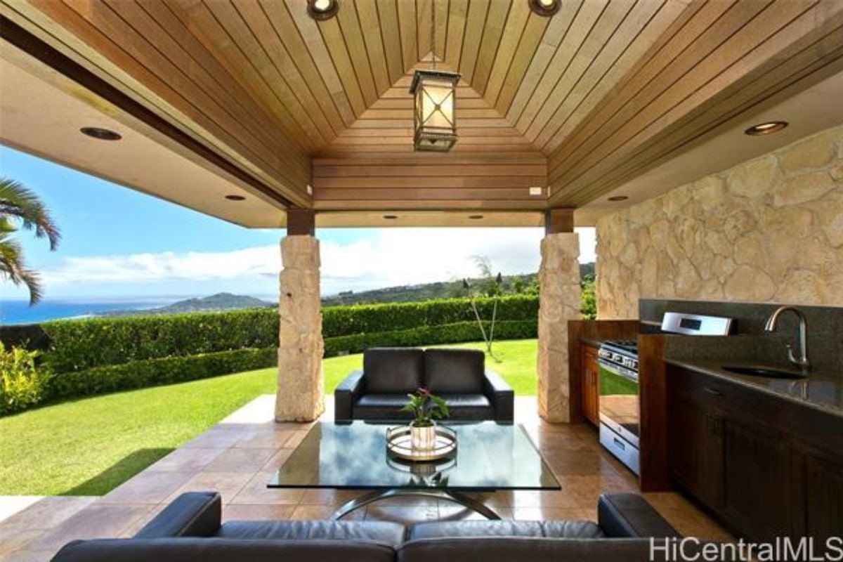 An outdoor living area with a vaulted ceiling, couches, a glass center table, and a kitchen.