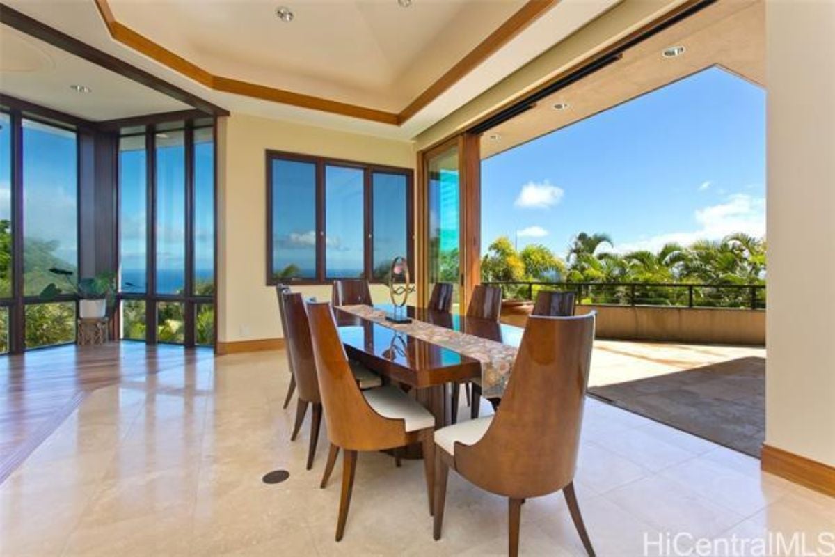 A dining room with windows, a tiled floor, and a long table surrounded by chairs.