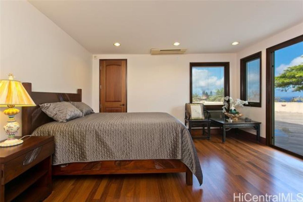 A bedroom with a bed, nightstand, windows, wooden floor, and a chair with a coffee table.