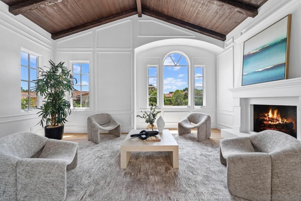 A living room with a vaulted ceiling, windows, a houseplant, a painting, a fireplace, a rug, chairs, and a center table.