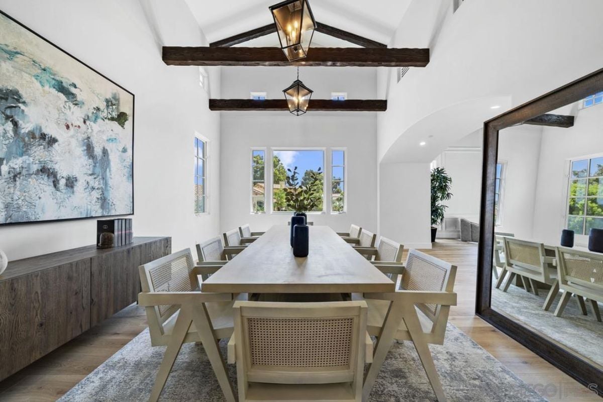A dining room with an exposed beam ceiling, a painting, a rug, and a long table surrounded by chairs.
