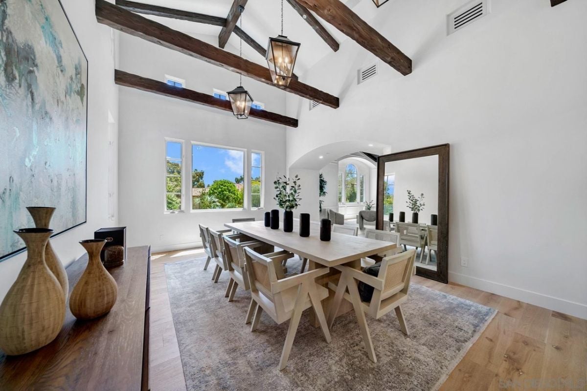 A dining room with an exposed beam ceiling, vases, a mirror, a painting, a rug, and a long table surrounded by chairs.