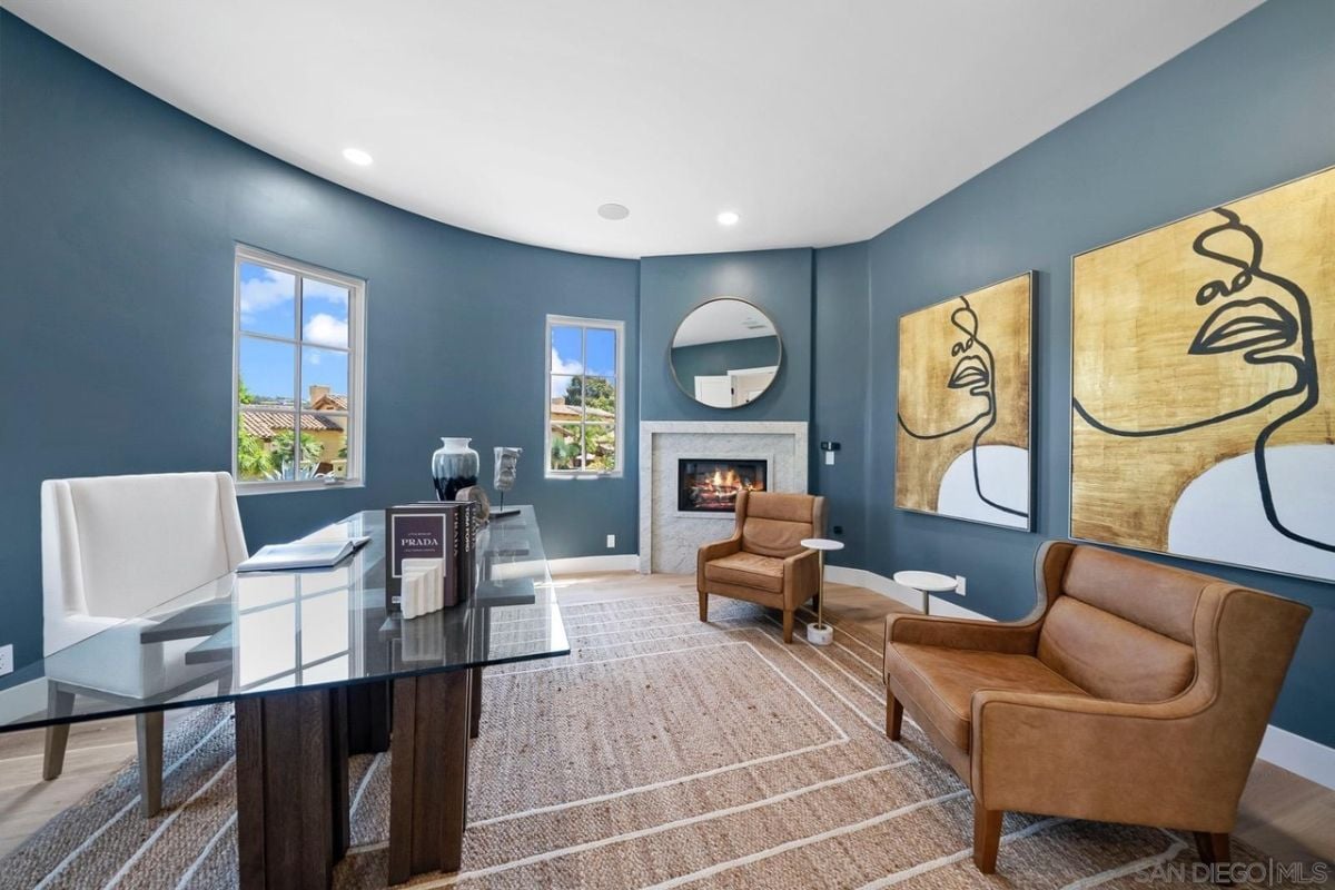 A home office with windows, paintings, a fireplace, and a glass desk with chairs.