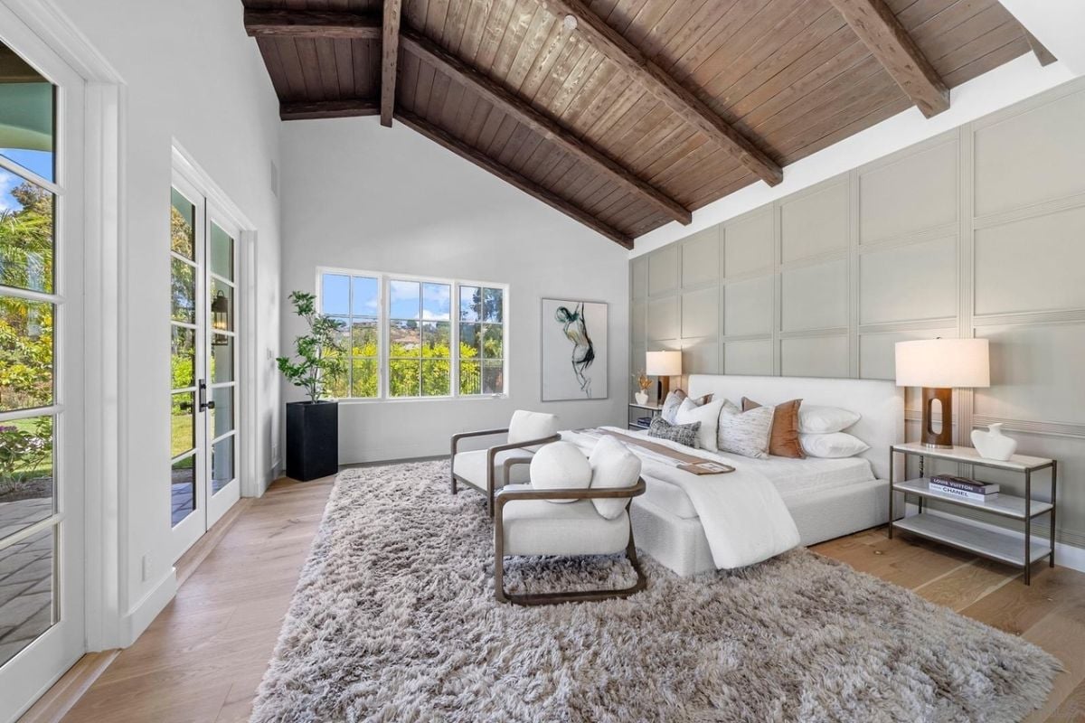 A bedroom with a bed, windows, an exposed beam ceiling, a painting, a rug, nightstands and chairs.