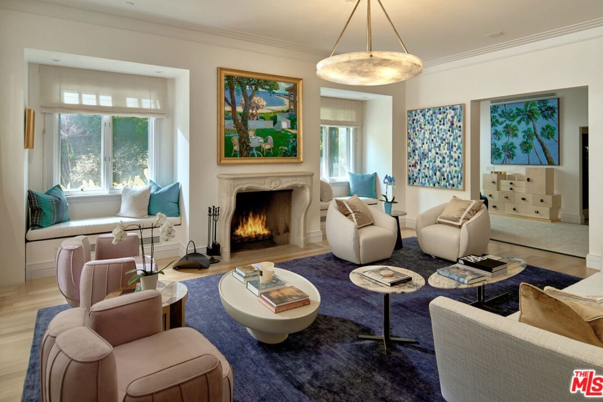 A living room with windows featuring window seating, paintings, a fireplace, a rug, chairs, a couch, and coffee tables.