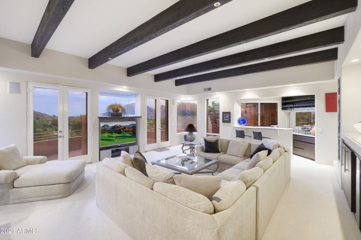 A living room with an exposed beam ceiling, windows, a TV, white couches, and a glass center table.