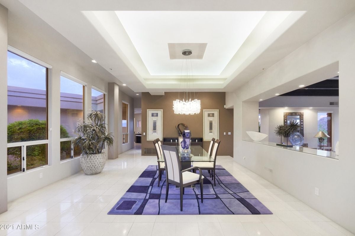 A dining room with windows, a vase, a chandelier, a rug, and a glass dining table surrounded by chairs.
