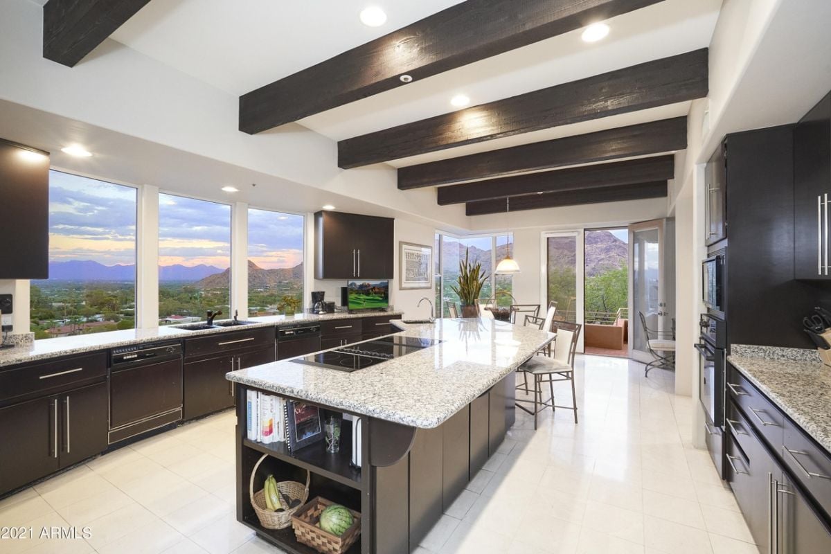 A kitchen with an exposed beam ceiling, windows, countertops, cabinets, and a sink.