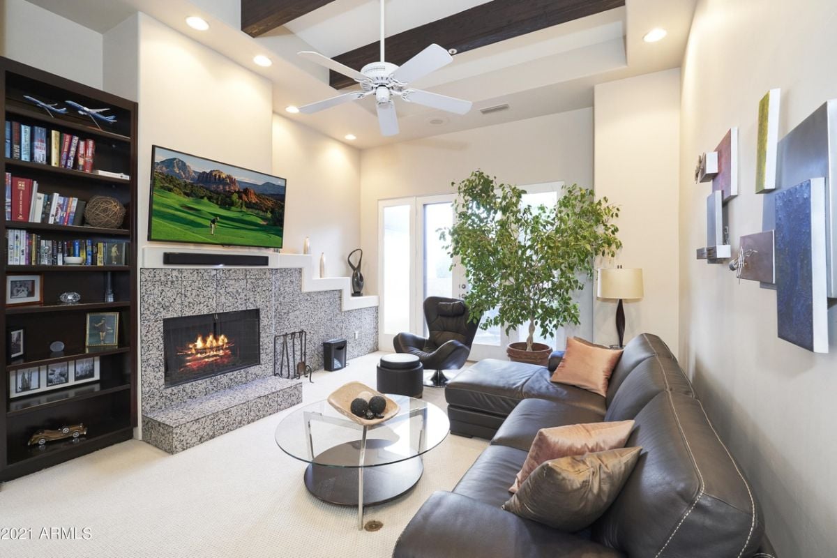 A family room with a shelf, a TV, a fireplace, a ceiling fan, a chair, a houseplant, a couch, and a round glass center table.