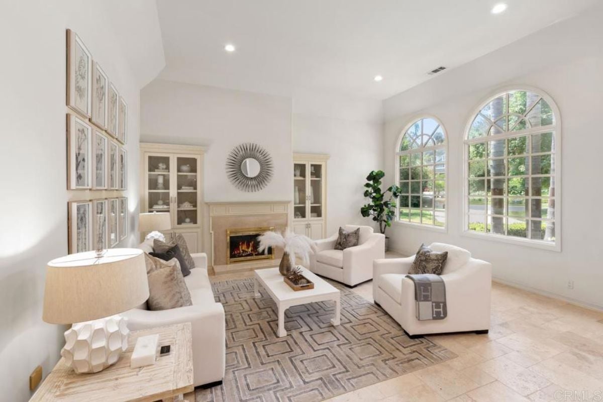 A living room with windows, a fireplace, paintings, a couch, chairs, and a center table.