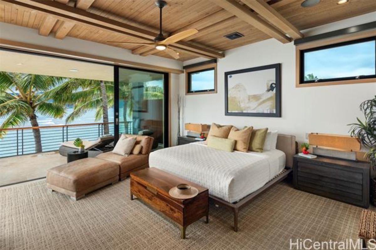 A bedroom with a bed, exposed beam ceiling, nightstands, comfy chair, and ceiling fan.