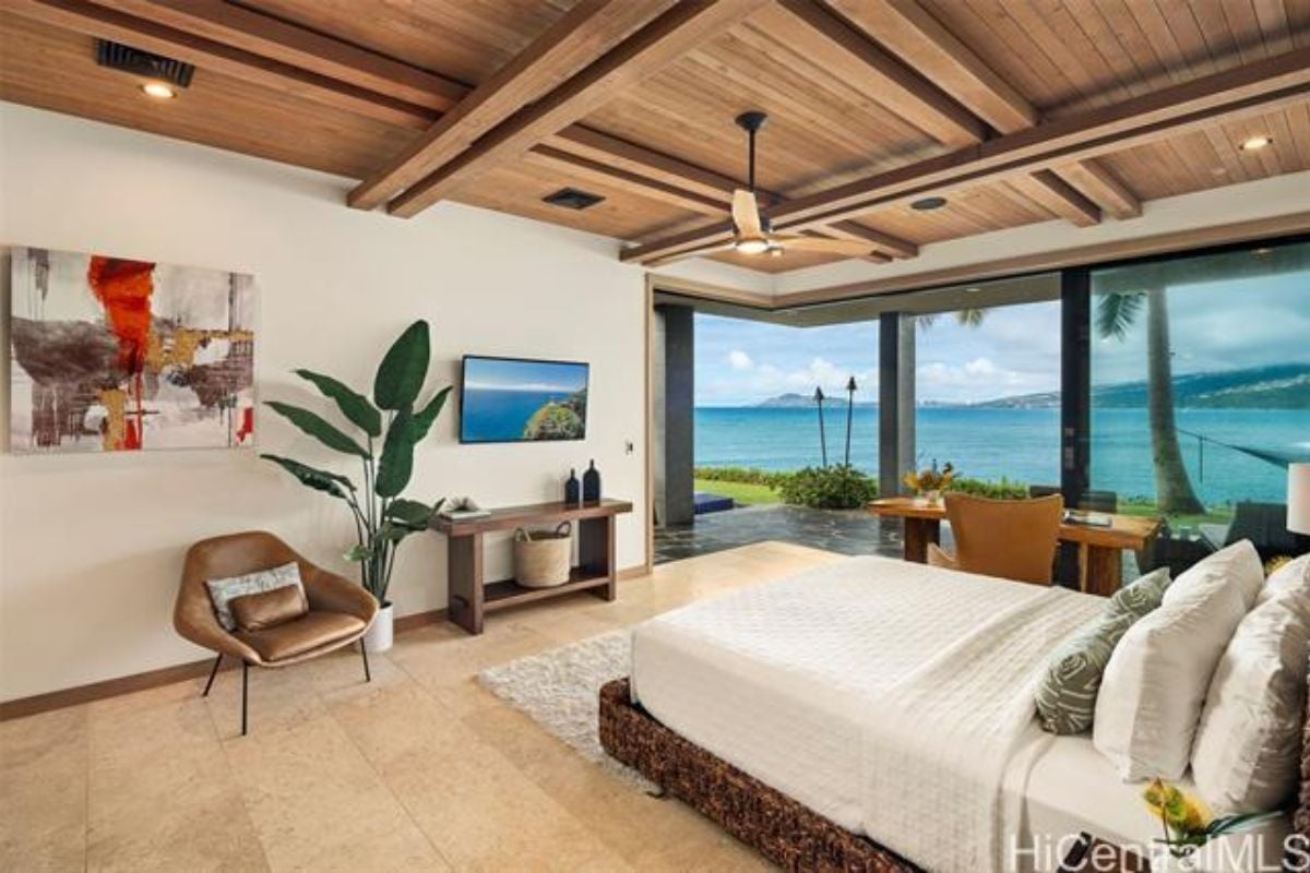 A bedroom with a bed, exposed beam ceiling, TV, chairs, desk and ceiling fan.