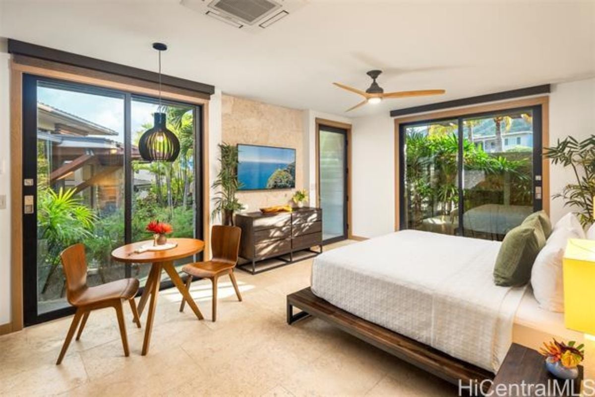 A bedroom with a bed, ceiling fan, TV, cabinet, and coffee table with chairs.