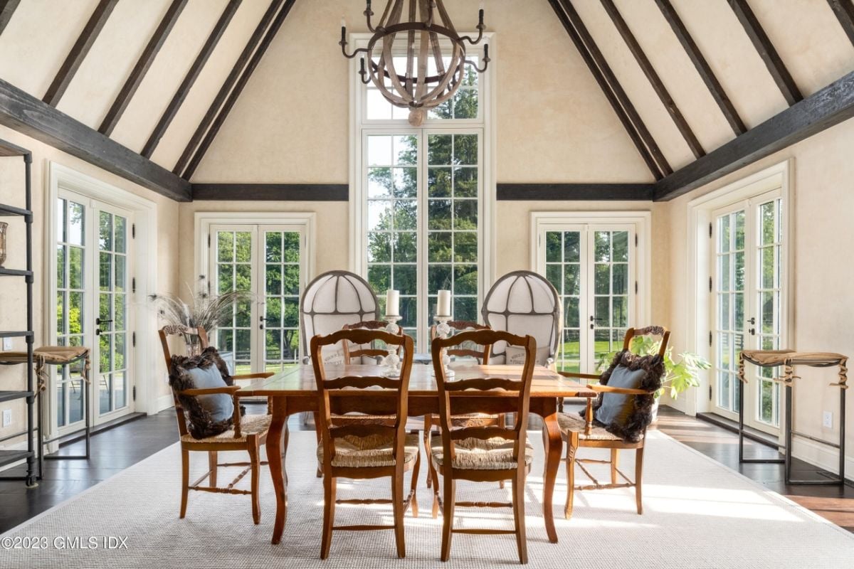 A dining room with a vaulted ceiling, chandelier, windows, a rug, and a dining table surrounded by chairs.