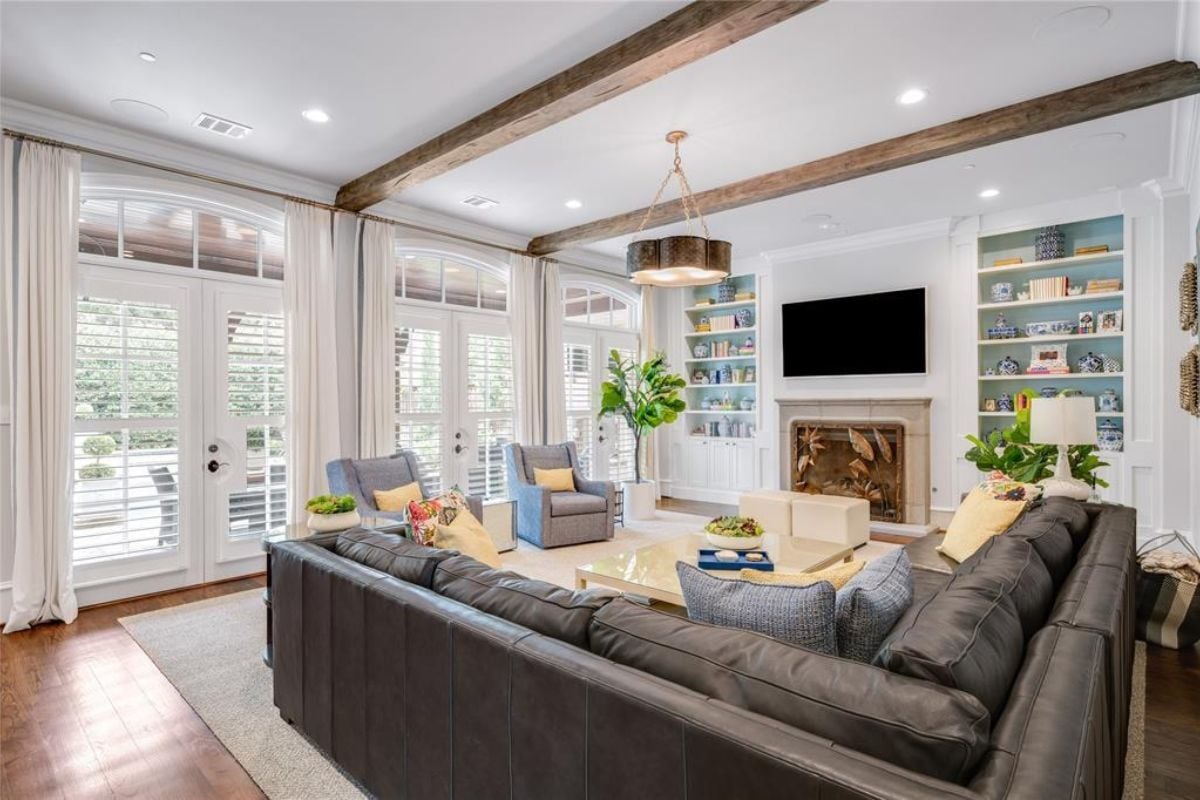 A living room with windows, an exposed beam ceiling, a TV, a fireplace, shelves, a couch, chairs, and a coffee table.