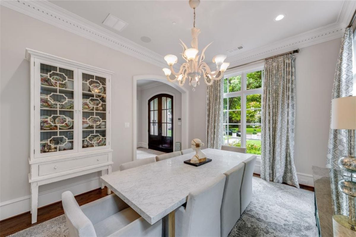 A dining room with a chandelier, a cabinet, windows, a rug, and a dining table surrounded by chairs.