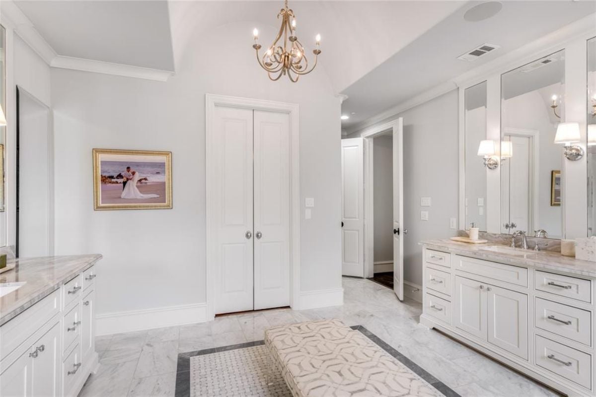 A bathroom with a vanity, a chandelier, a painting, cabinets, and a bench.
