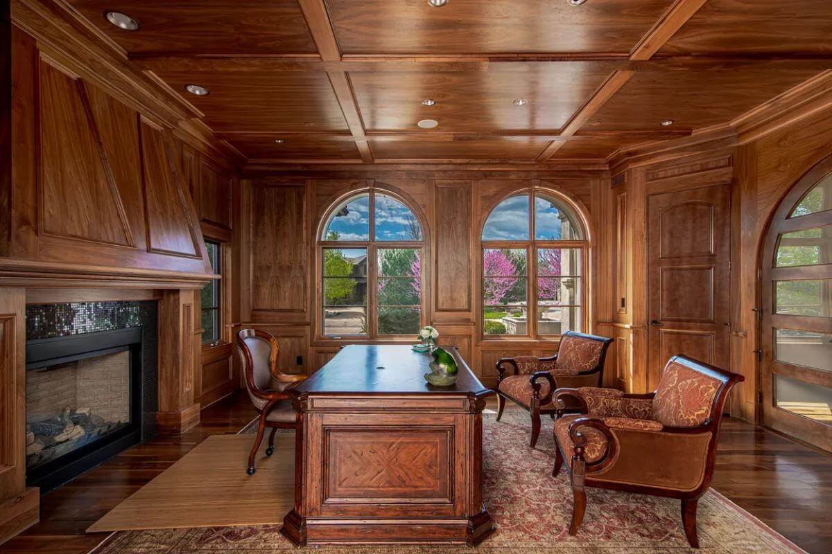 Office with a wooden desk, a fireplace, and wood-paneled walls that extend the the coffered ceiling.