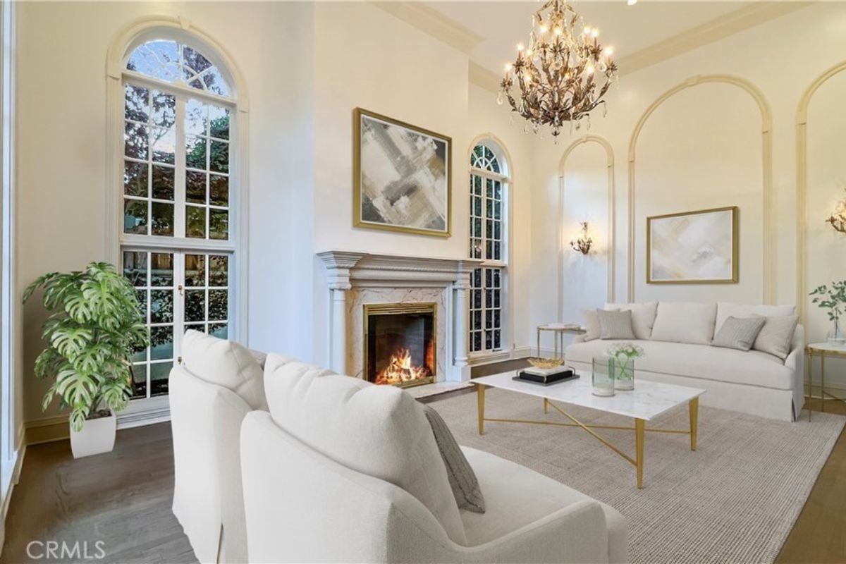 A living room with windows, a chandelier, paintings, a fireplace, a rug, couches, and a coffee table.