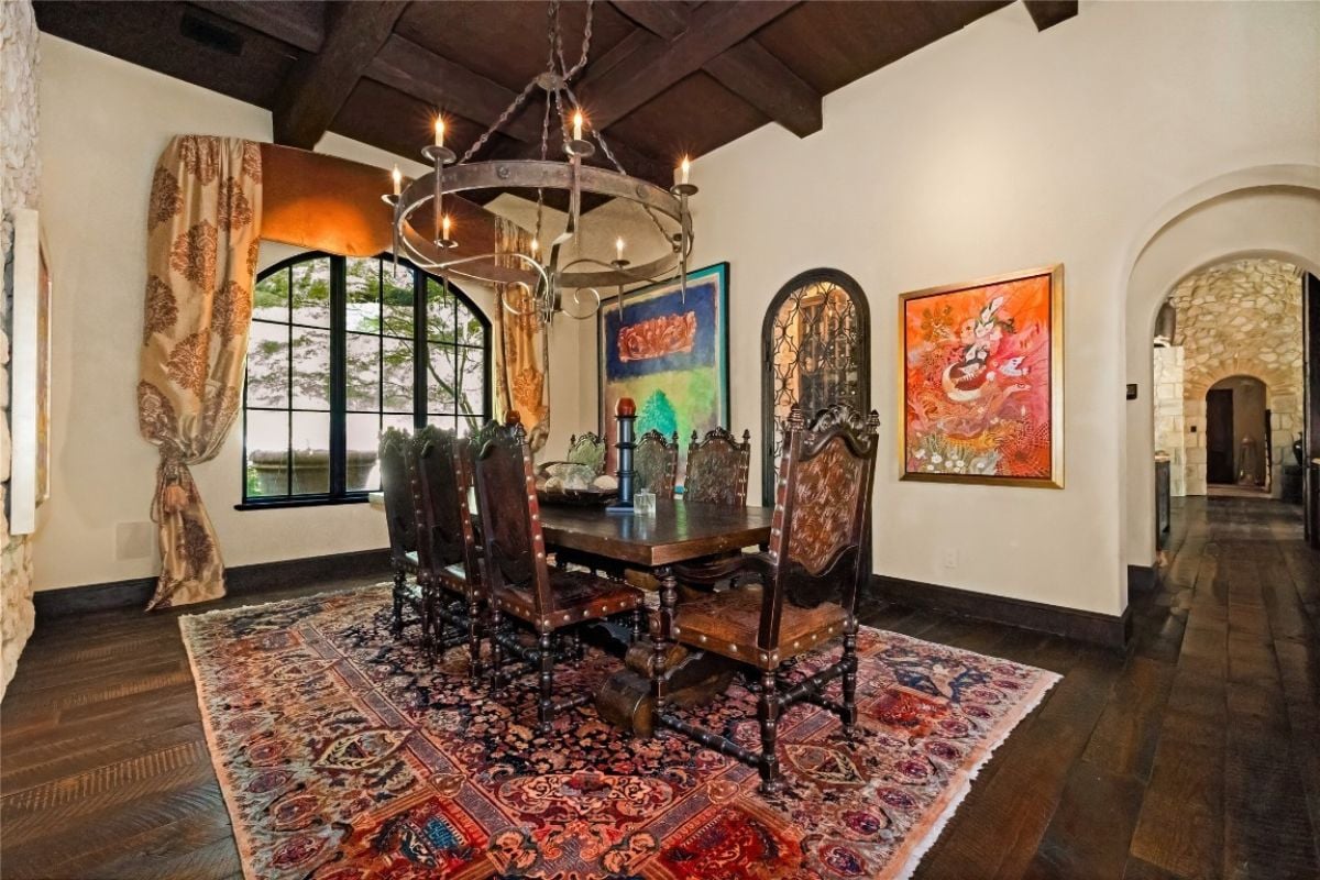 A dining room with an exposed beam ceiling, a window, a chandelier, paintings, a rug, and a dining table surrounded by chairs.
