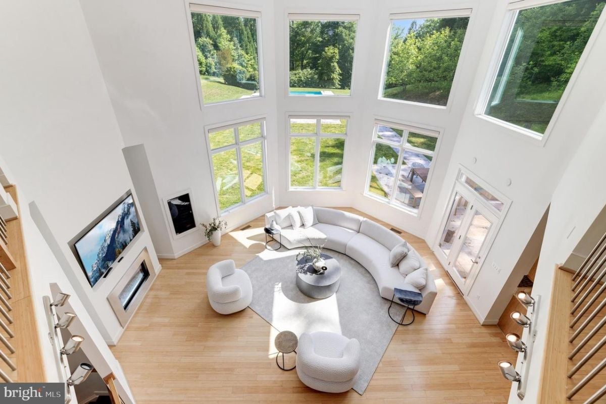 A living room with a high ceiling, windows, a TV, a fireplace, a curved couch, chairs, a rug, and a coffee table.