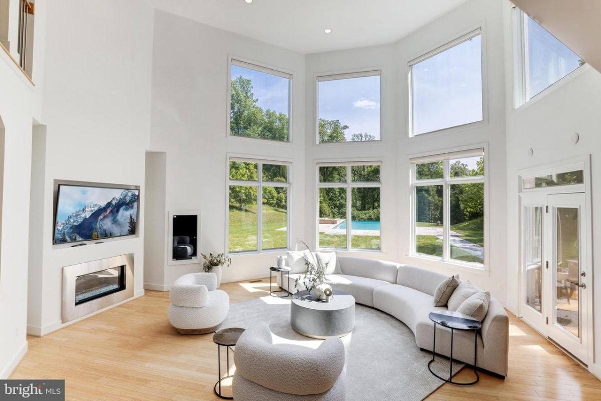 A living room with a high ceiling, windows, a TV, a fireplace, a curved couch, chairs, a rug, and a coffee table.