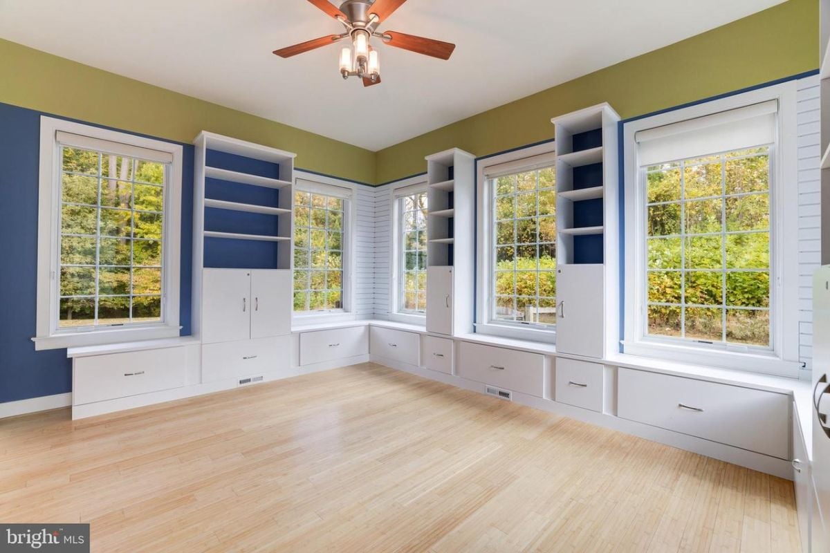 A bedroom with a bed, windows, a ceiling fan, cabinets, and drawers.