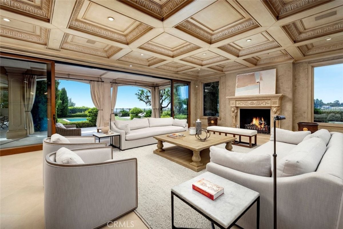 A living room with a ceiling with molding, a fireplace, a painting, windows, a rug, white couches, chairs, and a center table.