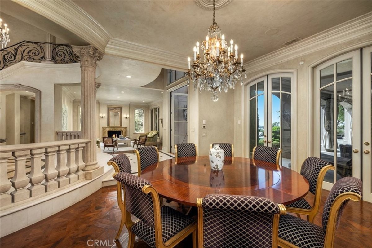 A dining room with a chandelier, windows, and a round table surrounded by chairs.