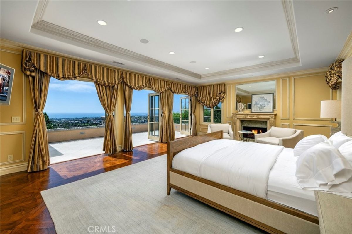 A bedroom with a bed, a rug, a fireplace, a painting, and glass doors with curtains leading to the balcony.