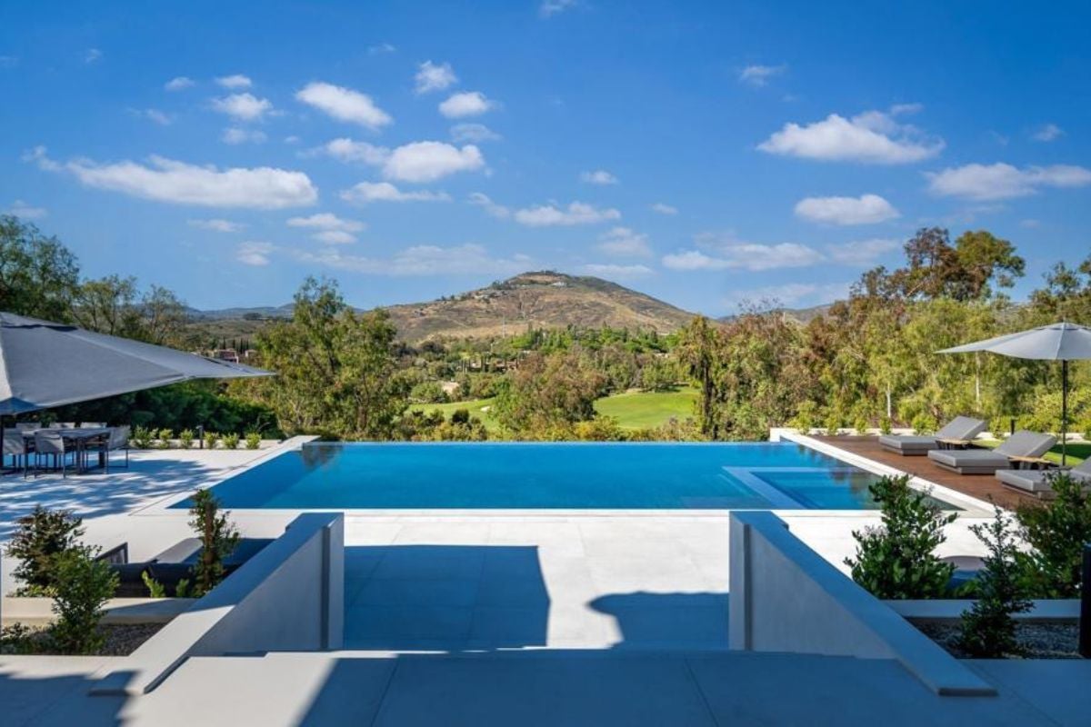A swimming pool with an umbrella and comfy chairs.