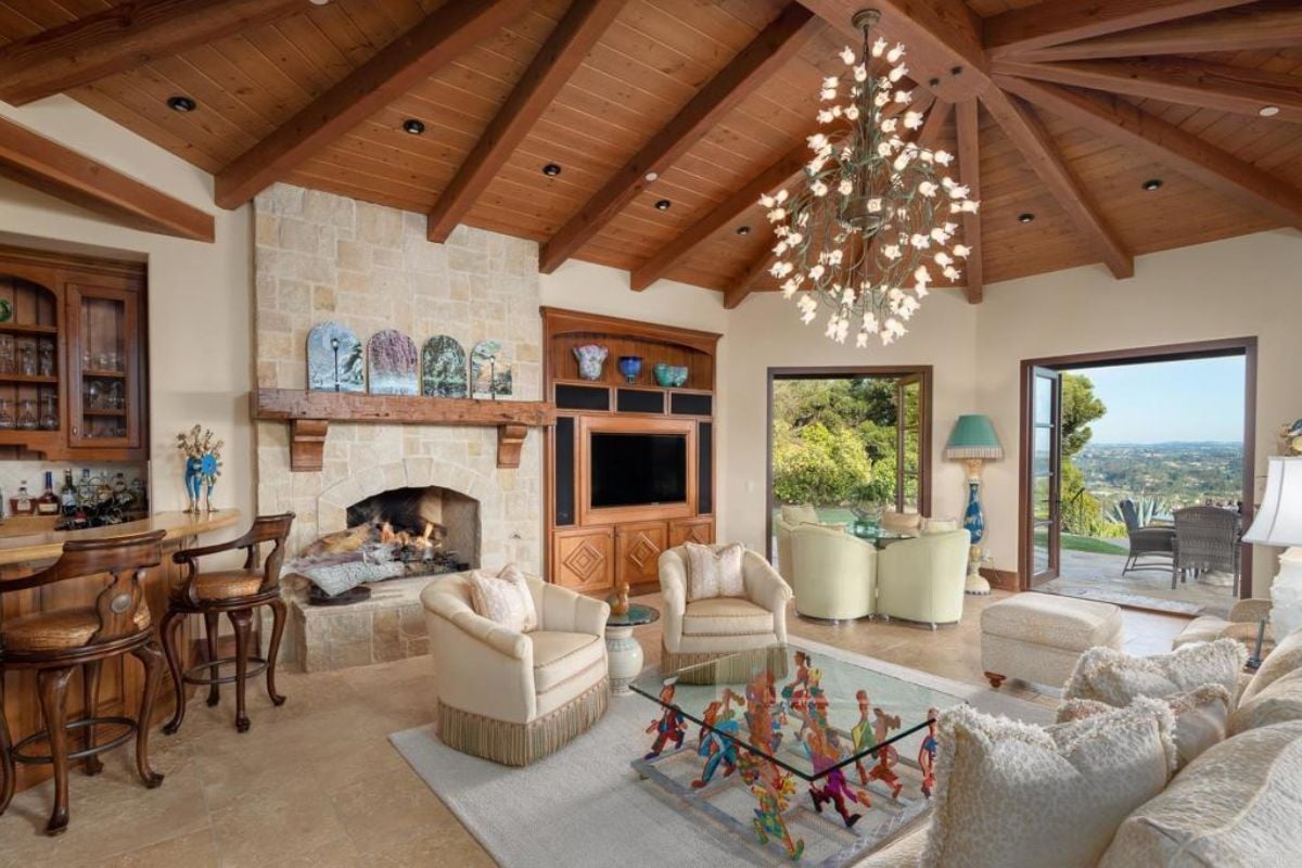 A living room with an exposed beam ceiling, a chandelier, a fireplace, a couch, a rug, a coffee table, and a bar.
