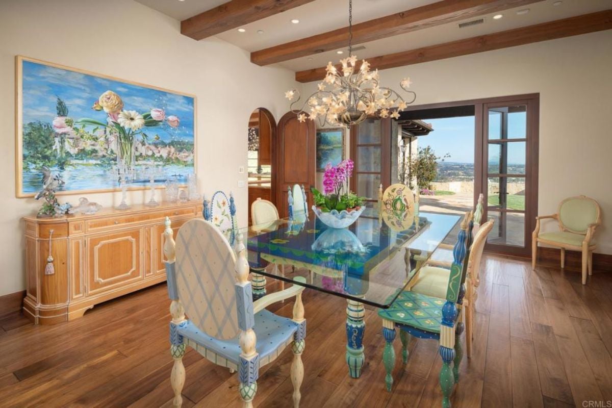 A dining room with a chandelier, an exposed beam ceiling, a painting, a cabinet, and a glass dining table surrounded by chairs.