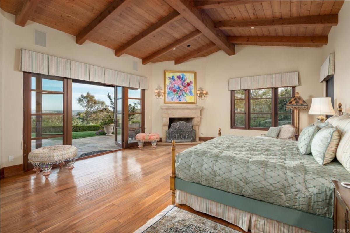 A bedroom with a bed, an exposed beam ceiling, a fireplace, a painting, and a window.