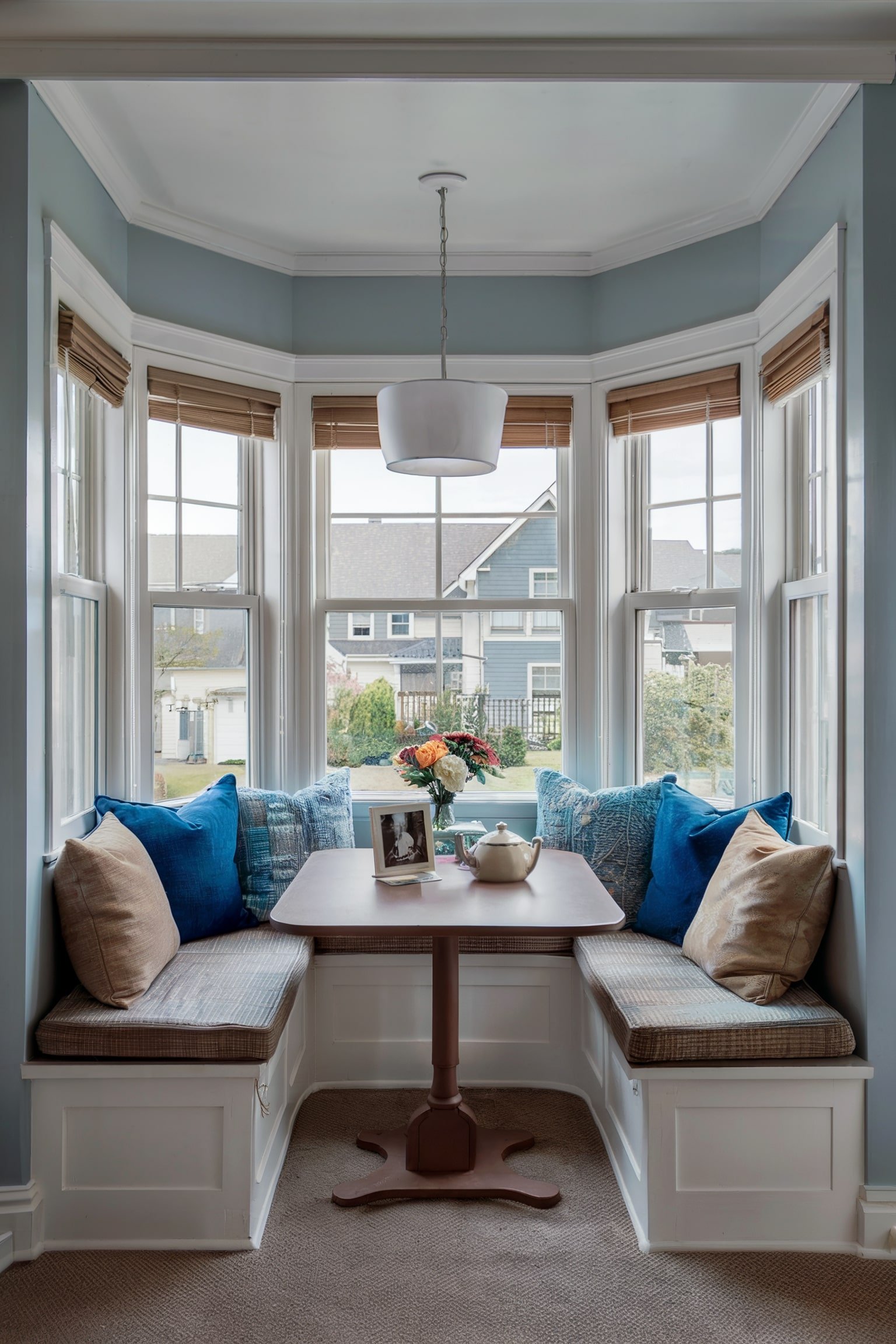Breakfast nook with built-in seating, cushions, and a table by a bay window overlooking houses.