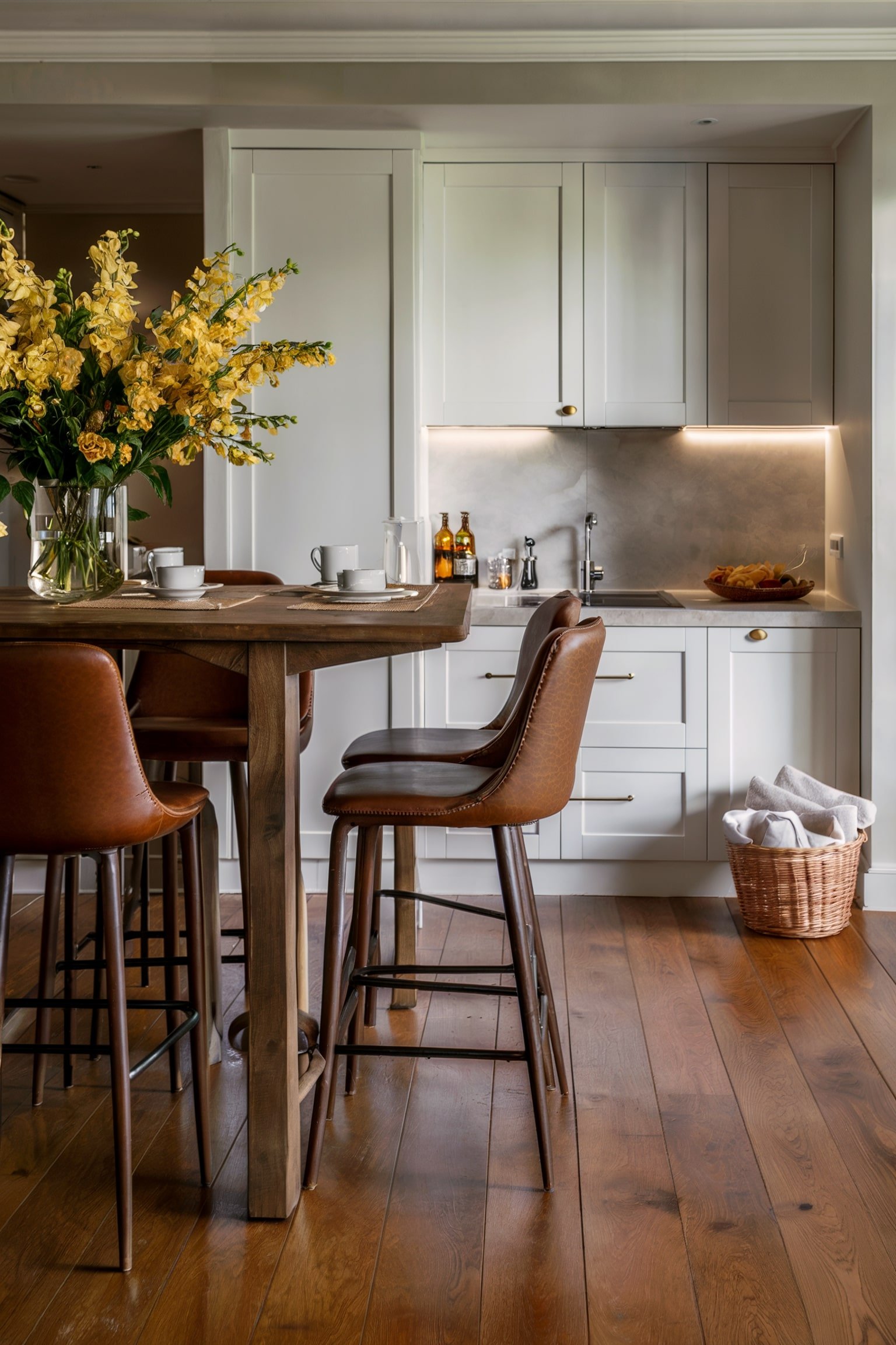 Kitchen with a high-top table, leather bar stools, white cabinets, and a vase of yellow flowers.