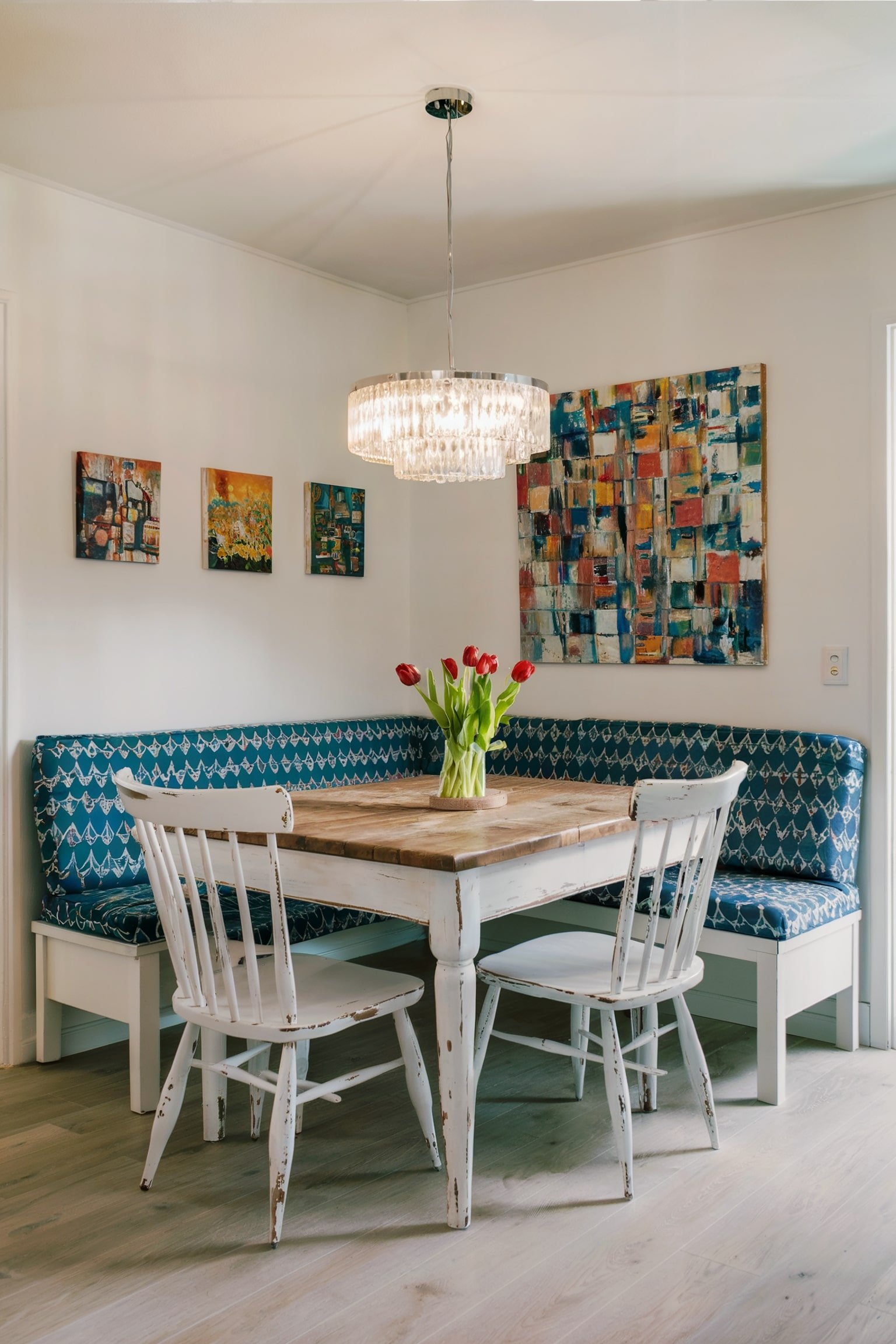 L-shaped blue bench, rustic table, white chairs, crystal chandelier, and colorful wall art.