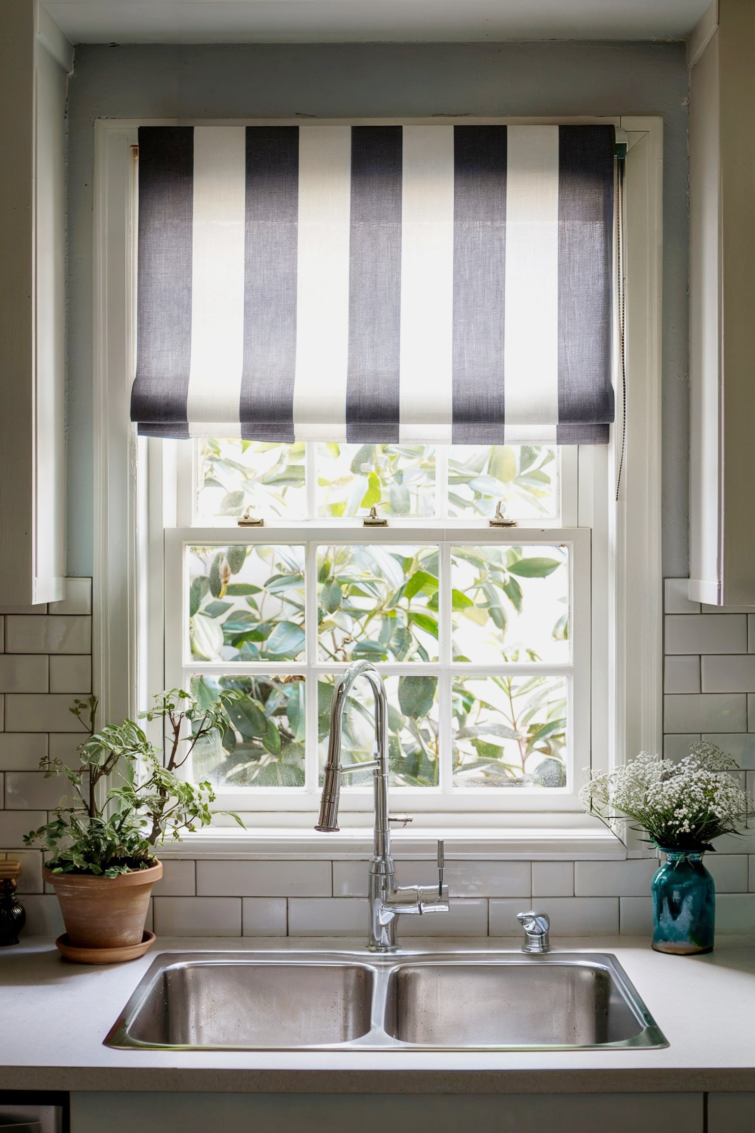 Kitchen window with a striped Roman shade, double sink, and potted plants on the counter.