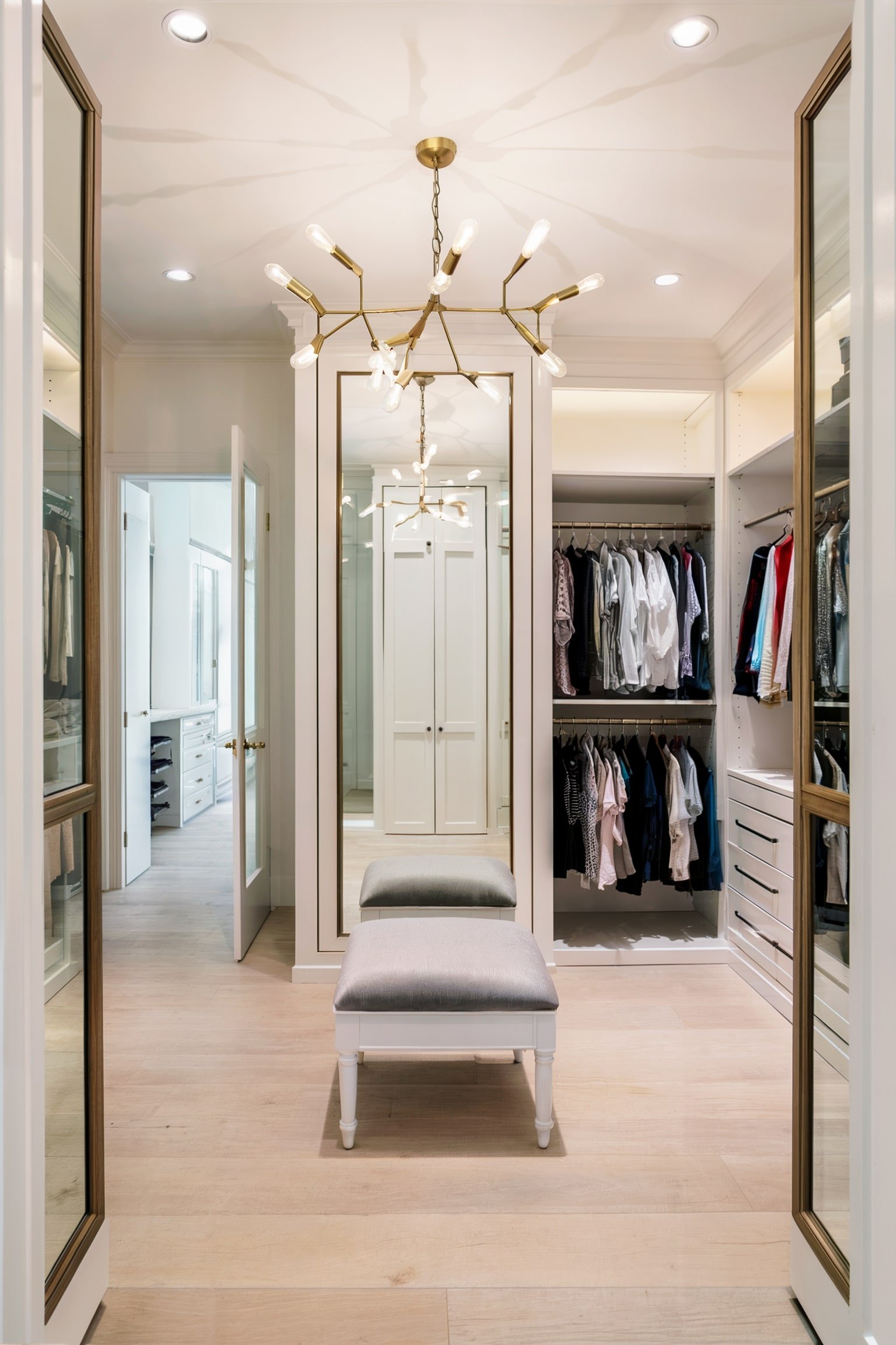 A bright, luxurious walk-in closet with mirrors, a bench, and a modern chandelier